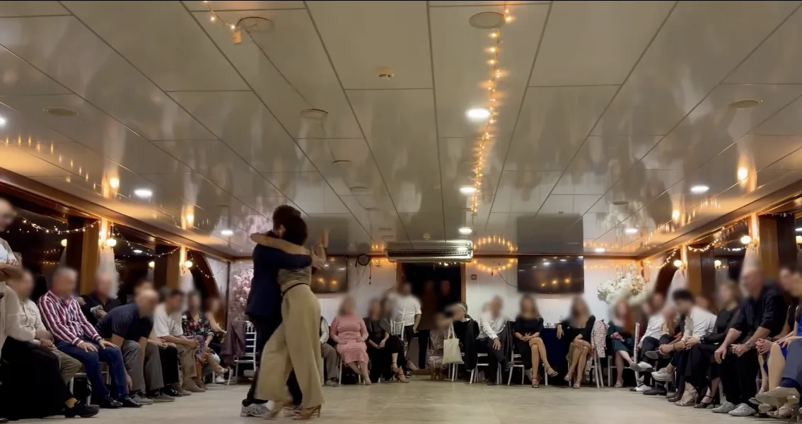 Emre Konuk and Majo Martirena dancing in close embrace on the floor of the Bosphorus boat milonga, with guests seated around the room under string lights