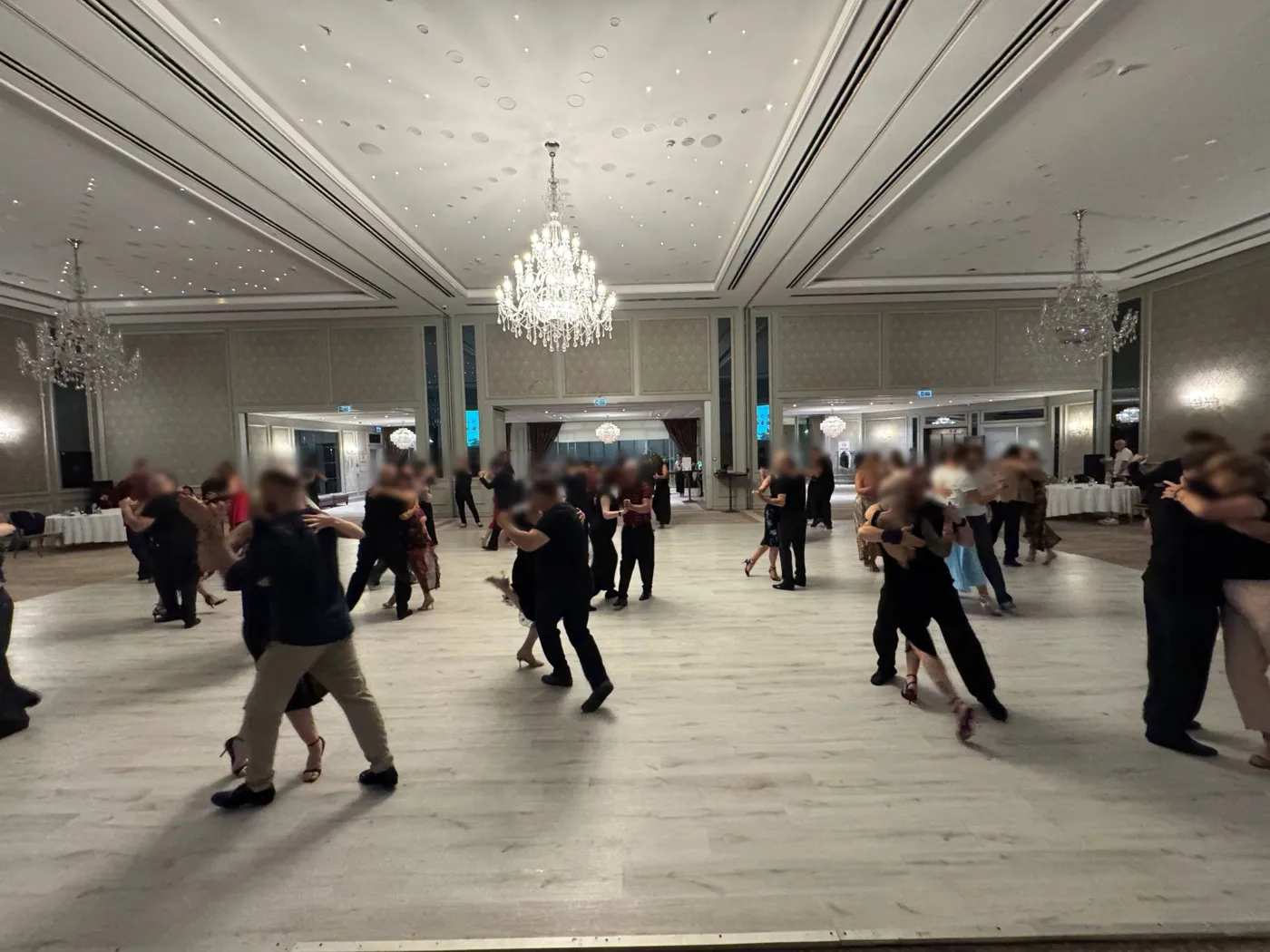 Couples dancing in close embrace on a pale wooden floor in a grand ballroom with crystal chandeliers