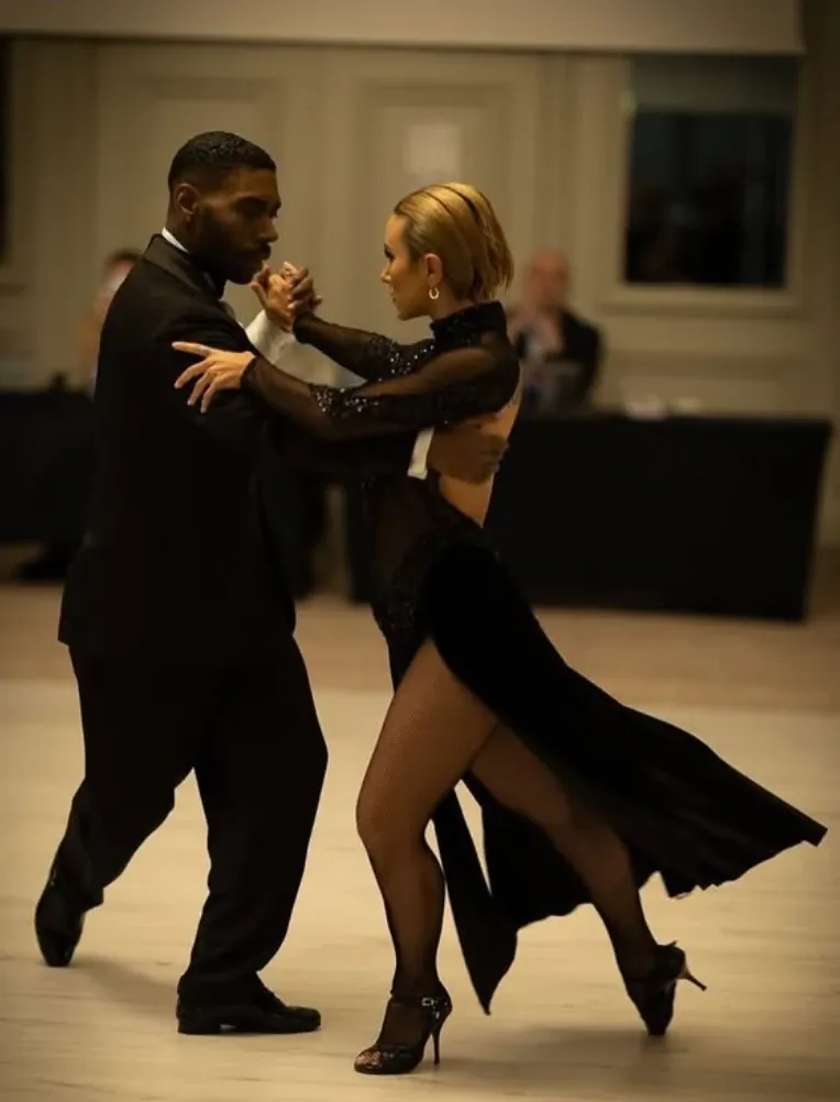 A Che Tango couple dancing close embrace in black outfits on a ballroom floor