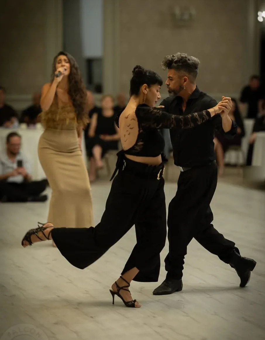 Rodrigo Fonti and Majo Martirena dancing in black outfits on a pale wooden floor while a singer performs behind them